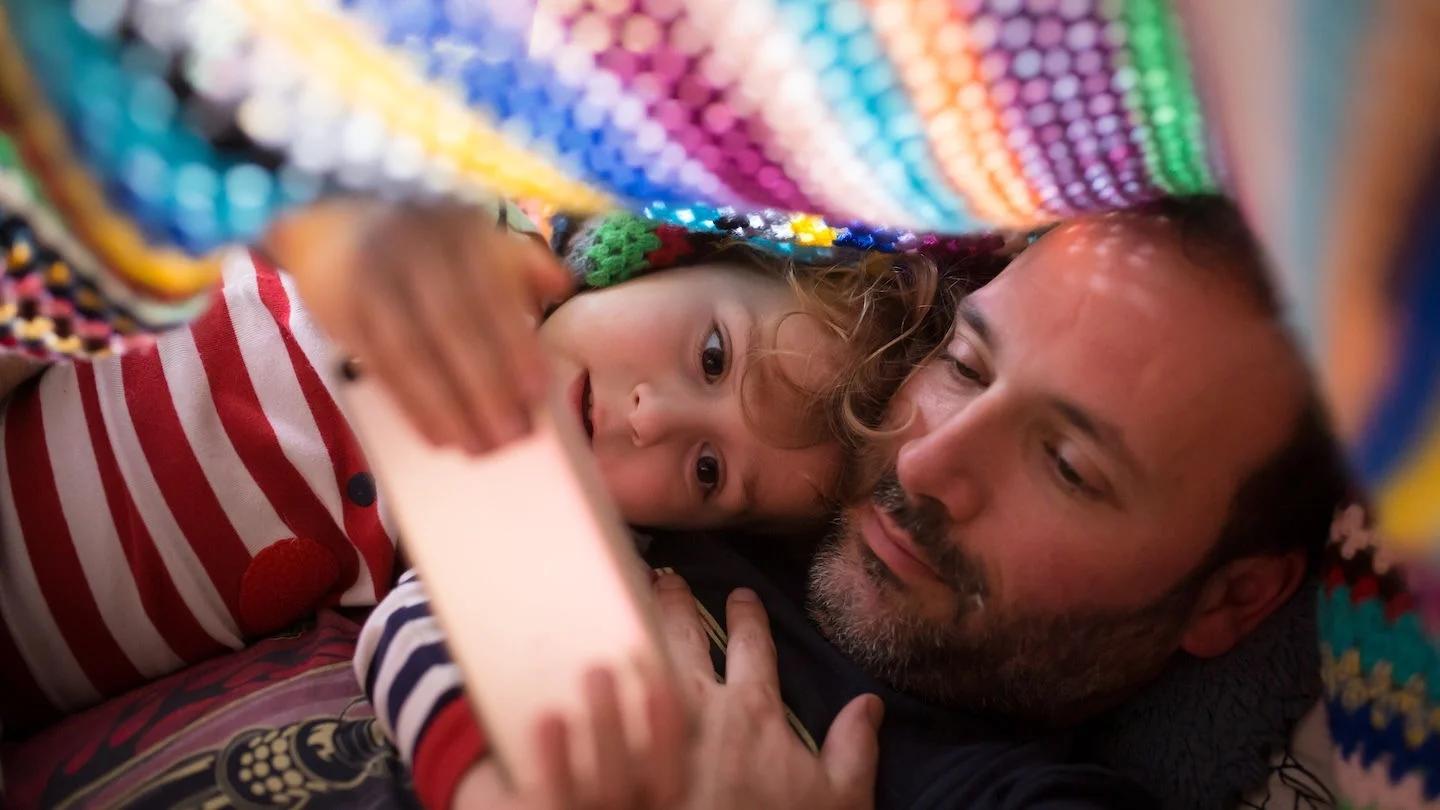 A dad and daughter under a blanket watching a video on a mobile device.