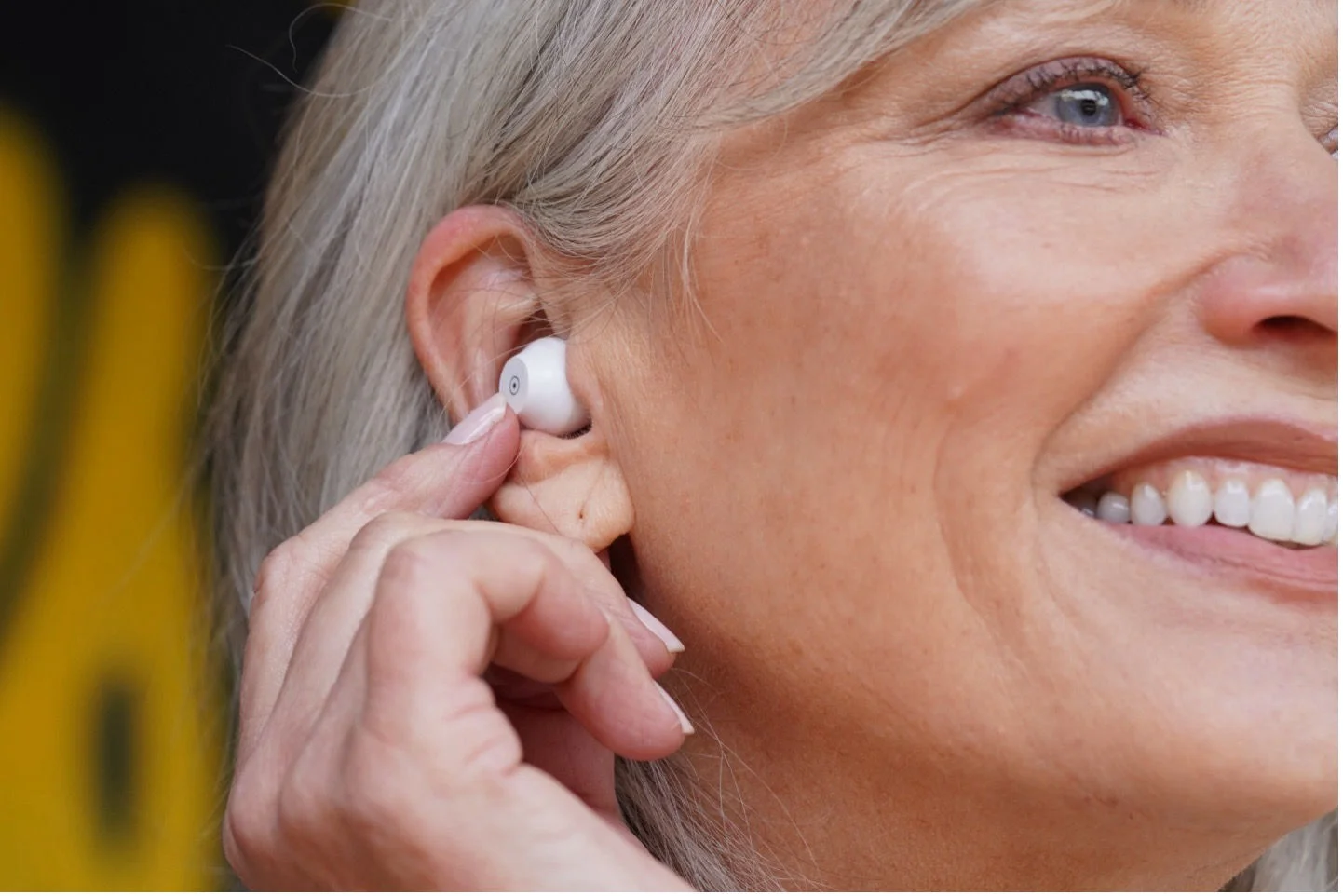 Close-up of a smiling woman inserting a white wireless earbud into her ear.